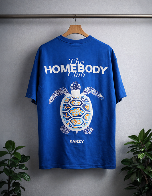 Blue t-shirt with turtle graphic and 'The Homebody Club' text on a hanger against a gray background.